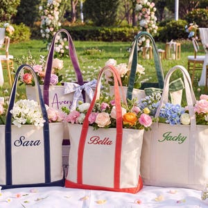 May include: Four canvas tote bags in cream with colored trim and handles. Each bag features an embroidered name: Sara in navy, Avery in purple, Bella in red, and Jacky in green. The bags are filled with assorted colorful flowers and greenery.