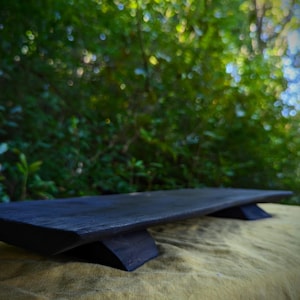 May include: A dark wooden serving board with a rectangular shape and two supporting legs. The board has a gently curved top and a dark, matte finish. The background shows blurred green foliage.