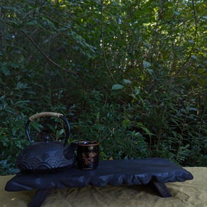 May include: A black cast iron teapot with a woven handle rests on a dark wooden table, alongside a small brown teacup. The teapot features a decorative wave pattern. The table is on a yellow-green cloth, with a backdrop of dense green foliage.