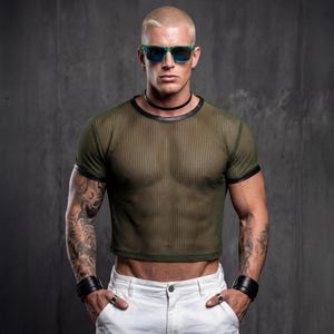 May include: Olive green mesh crop top with black trim, paired with white pants. The model is wearing green sunglasses, a black choker, and black wrist cuffs. The background is a dark gray wall.