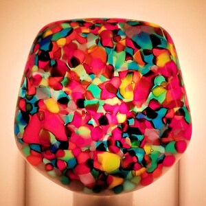 May include: A colorful nightlight with a multi-colored glass shade plugged into a standard electrical outlet. The glass shade has a mosaic pattern of red, yellow, blue, green, and black. The outlet has a sticker that says "SKU 516".