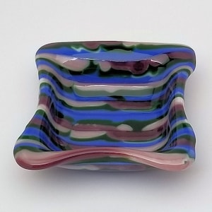 May include: A small, square-shaped glass dish with a wavy, rippled design. The dish is made of layered glass in shades of blue, green, pink, and white.