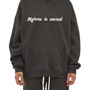 Peut inclure: Un sweat à capuche gris foncé avec le texte "Nature is sacred" en lettres cursives blanches.