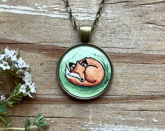 May include: A necklace with a round pendant featuring a hand-painted watercolor illustration of a fox curled up asleep on a green background.