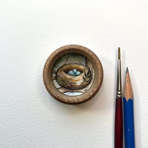 Puede incluir: Un pequeño marco redondo de madera con una pintura de acuarela de un nido de pájaro con tres huevos azules en su interior. El nido está hecho de ramitas y está rodeado de hojas verdes. La pintura está rodeada por un borde blanco.