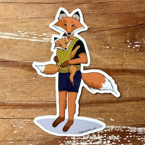 May include: A sticker featuring a cartoon fox holding a baby fox. The adult fox wears a black shirt, blue shorts, and a yellow polka-dot carrier. The sticker has a white border and the artist's website is visible.