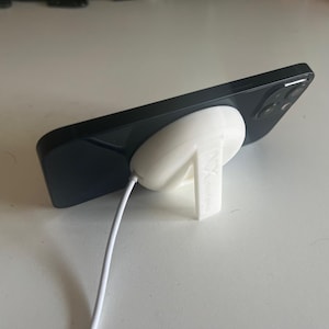 Puede incluir: Un soporte de teléfono blanco impreso en 3D con un teléfono inteligente negro descansando en la parte superior. El soporte tiene una base circular y un solo soporte vertical. Un cable de carga blanco está conectado al teléfono.
