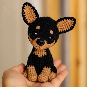 Può includere: Un cane Chihuahua all'uncinetto fatto a mano in nero e marrone chiaro. Il cane amigurumi ha grandi occhi espressivi ed è seduto in posizione eretta. Le orecchie, le zampe e il muso sono marroni, mentre il corpo e la testa sono neri. È un giocattolo carino e coccoloso.
