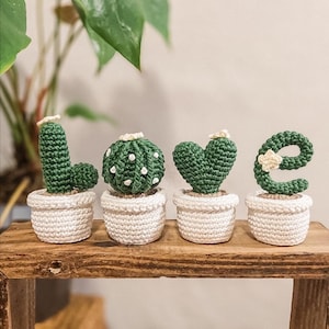 Peut inclure: Quatre petits cactus en crochet dans des pots blancs qui épellent le mot "LOVE". Les cactus sont verts et ont des accents blancs. Les pots sont blancs et ont une texture en crochet.