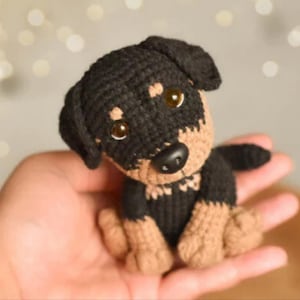 Puede incluir: Un juguete de perro hecho a mano con ganchillo, negro y marrón claro. El pequeño animal de peluche tiene una cabeza y un cuerpo negros con patas y hocico de color marrón claro. El perro tiene ojos grandes, redondos, color ámbar y una nariz negra. El juguete está en una mano.