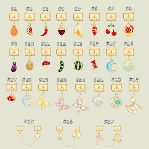 Puede incluir: Una colección de colgantes con dijes dorados con varios diseños, incluyendo frutas, verduras y formas caprichosas. Los dijes están dispuestos en filas, mostrando una variedad de colores y estilos, perfectos para la fabricación de joyas o manualidades.