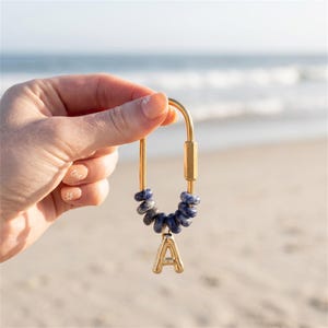 Puede incluir: Un llavero de mosquetón de metal dorado con una serie de pequeñas cuentas redondas de piedra azul y gris jaspeada ensartadas en la curva inferior, con un dije de letra A de color dorado colgando del centro de las cuentas.