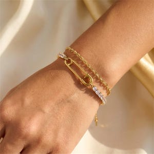 Puede incluir: Pulseras doradas apiladas en una muñeca. Una pulsera presenta un diseño de imperdible, y otra es una cadena delicada. La tercera pulsera está adornada con piedras rectangulares transparentes. Las pulseras se muestran sobre un fondo claro.