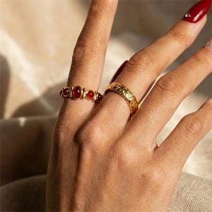 Puede incluir: Dos anillos de tono dorado en los dedos. Un anillo presenta una serie de pequeñas piedras rojas ovaladas y lisas dispuestas en fila. El segundo anillo tiene una superficie texturizada y grabada con un patrón repetitivo en toda la banda dorada.