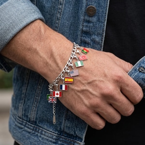 Könnte beinhalten: Ein silbernes Armband mit Miniatur-Flaggen-Anhängern. Die Flaggen repräsentieren verschiedene Länder, darunter Deutschland, Frankreich und Spanien. Das Armband wird am Handgelenk getragen, mit einer Jeansjacke im Hintergrund.