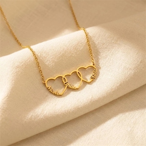 Puede incluir: Un collar de tono dorado con tres colgantes entrelazados en forma de corazón. Cada corazón tiene grabado un nombre en letra cursiva, específicamente Nicole, Ethan y Olivia, unidos por una delicada cadena de eslabones dorados.