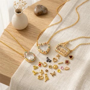 Puede incluir: Tres collares dorados con colgantes transparentes, incluyendo una lágrima, un corazón y un marco rectangular con la palabra MISS en su interior. Cada colgante presenta un borde de piedras multicolores. Se incluye una colección de pequeños dijes dorados.