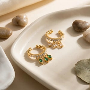 Puede incluir: Dos pendientes tipo ear cuff dorados con una hilera de piedras transparentes pavé a lo largo de la banda. Uno presenta tres piedras transparentes colgantes en talla baguette, y el otro tres piedras verdes colgantes en talla marquesa.