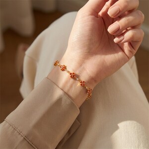 Puede incluir: Un brazalete dorado con dijes rojos en forma de flor y pequeñas gemas blancas. El brazalete se lleva en una muñeca, mostrando su delicado diseño y detalles intrincados. Es una pieza de joyería.