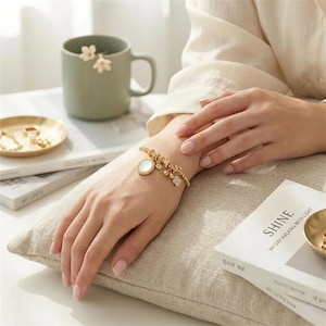 Puede incluir: Una pulsera dorada con cuentas, un colgante redondo blanco y pequeños abalorios se lleva en la muñeca. La pulsera está en el brazo de una persona, apoyada en una almohada beige. Un plato dorado y una taza verde están en el fondo.