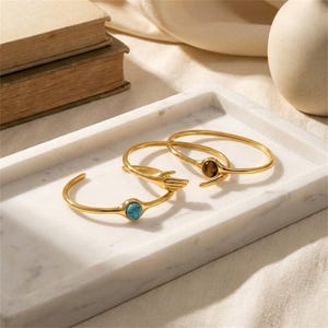 Puede incluir: Dos pulseras rígidas de metal dorado. Una pulsera presenta una piedra redonda de color turquesa. La segunda pulsera muestra un motivo de mano esculpida que sostiene una piedra redonda de ojo de tigre marrón. Ambas piezas tienen acabado pulido.
