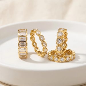 May include: A set of four gold-toned rings featuring clear stones. One ring has emerald-cut stones, one has marquise-cut stones, one has heart-shaped stones, and one has round-cut stones. Each ring is crafted with a metallic gold-colored band.