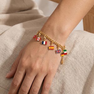Op de afbeelding: Goudkleurige bedelarmband met een kettingontwerp. De armband is versierd met kleurrijke emaille bedels die verschillende nationale vlaggen vertegenwoordigen, waaronder Canada, Frankrijk, Spanje, het Verenigd Koninkrijk en Duitsland. De armband wordt om de pols gedragen.