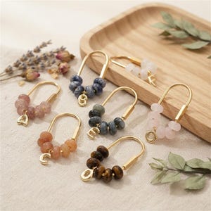 Pode incluir: Uma coleção de seis alfinetes em forma de U de metal dourado, cada um adornado com um grupo de pequenas contas redondas de pedras preciosas multicoloridas e um pingente dourado em forma de letra ou coração pendurado na base curva.
