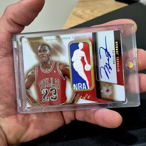 May include: A Michael Jordan Ultimate Signatures trading card encased in a clear plastic holder. The card features a photograph of Jordan in a red Bulls jersey, an embroidered NBA logo patch, a blue ink autograph, and the text 1 of 1.