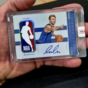 May include: A Luka Dončić National Treasures basketball trading card encased in a clear plastic holder. The card features a photo of the player in a blue Dallas Mavericks jersey, an NBA logo patch, and a blue ink autograph. Text includes DALLAS, MAVERICKS, and LUKA DONČIĆ.
