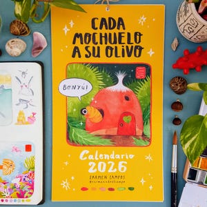 Puede incluir: Calendario amarillo de 2026 con el texto "Cada Mochuelo A Su Olivo" y una ilustración de una casa roja con una puerta verde en forma de corazón. El calendario también incluye el nombre "Carmen Campos" y el Instagram @carmenadestiempo.