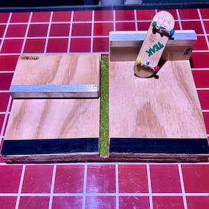 May include: A miniature wooden skateboard ramp set with a fingerboard. The set includes two wooden platforms with metal rails and a strip of green turf. A small skateboard with the word "TEAK" is on the ramp.