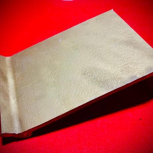 May include: A gold metal stand with a flat surface and a raised edge. The stand is angled and has a smooth, metallic finish.