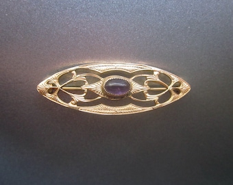 May include: An oval-shaped gold brooch featuring an openwork metal design with engraved leaf patterns. A single, smooth, oval-cut purple amethyst gemstone is set securely in the center of the piece, held by a delicate metal bezel setting.