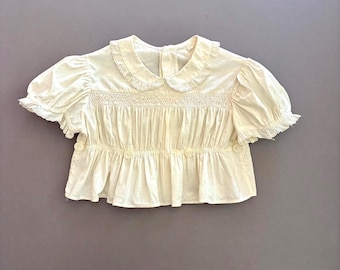 May include: A cream-coloured short-sleeve blouse featuring a rounded collar with lace trim, smocked detailing across the chest, puffed sleeves with ruffled edges, and a gathered waistline accented with four small buttons along the front.