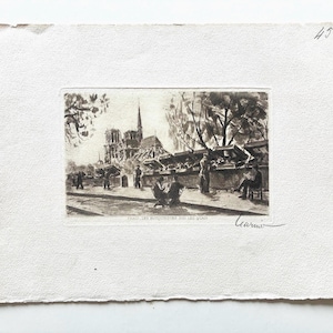 May include: A black and white etching on textured paper depicting a Parisian street scene with the Notre Dame cathedral in the background, bookstalls along the riverbank, and figures sitting at tables. The text reads PARIS, LES BOUQUINISTES DES QUAIS.