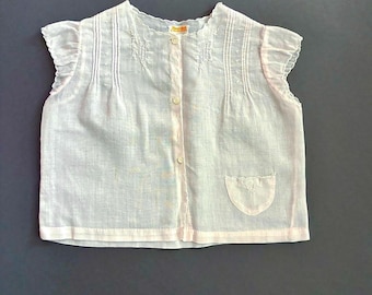 May include: A light pink, short-sleeved blouse featuring a button-down front, delicate pintuck detailing on the chest, and a small patch pocket. The garment has a scalloped neckline and ruffled cap sleeves, with a label visible at the collar.