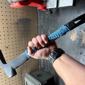 May include: A grey crowbar with a black end and a blue wrapped handle. The tool has a hammer head on one end and a forked end on the other. The handle is wrapped in blue cord.