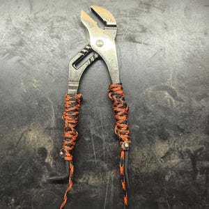 Puede incluir: Alicates de plata con paracord negro y naranja envuelto alrededor de los mangos.