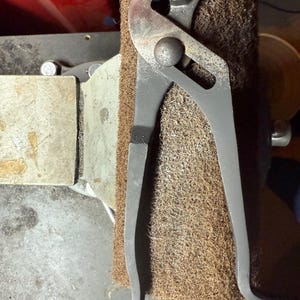May include: A pair of grey metal pliers with a textured grip, resting on a brown abrasive pad. The pliers have a curved handle and a toothed jaw, designed for gripping and turning objects. The tool is likely used for mechanical or construction tasks.