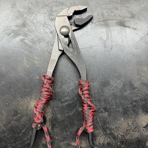 Può includere: Pinza a becco curvo con finitura argentata, avvolta da paracord nero e rosso. La pinza ha una ganascia curva e una punta affilata all'estremità.