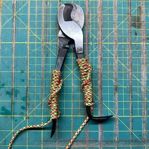 May include: A pair of black wire cutters with orange and green paracord wrapped around the handles. The cutters are on a green cutting mat.
