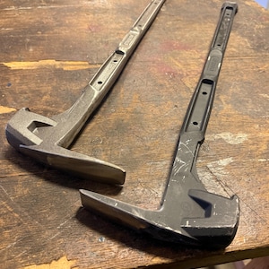 May include: Two metal pry bars, one silver and one black, rest on a weathered wooden surface. The tools have a claw at one end and a beveled edge at the other, designed for prying and demolition tasks. The silver bar shows signs of use.