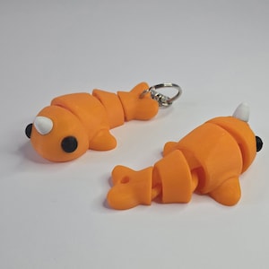 May include: Two orange articulated narwhal keychain toys. One narwhal has a silver key ring attached. Both narwhals have black eyes and white tusks.
