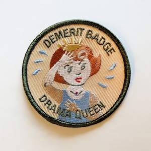 May include: A beige embroidered patch with a green border. The patch features a cartoon woman with red hair wearing a blue dress and a gold crown. The text "Demerit Badge Drama Queen" is embroidered around the woman.