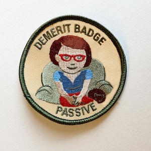 May include: A green embroidered patch with the text "Demerit Badge Passive" and a cartoon illustration of a person wearing red glasses and a blue shirt sitting in a chair.
