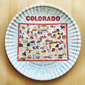 May include: A round, white tray with a ruffled edge and a map of Colorado. The map is in the center, surrounded by a light blue border with illustrations. The word "COLORADO" is in large red letters at the top.