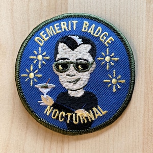 May include: A blue and green embroidered patch with the text "Demerit Badge Nocturnal" and a cartoon image of a person wearing sunglasses and holding a cocktail.