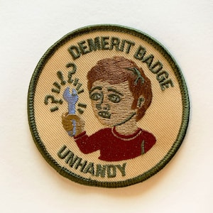 May include: A beige and green embroidered patch with a cartoon illustration of a person holding a wrench. The text on the patch reads "Demerit Badge Unhandy" and "?" symbols are around the wrench.