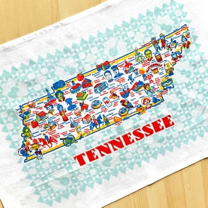 May include: A white tea towel with a blue and white geometric pattern. The center of the towel features a colorful map of Tennessee with the word "Tennessee" in red letters.